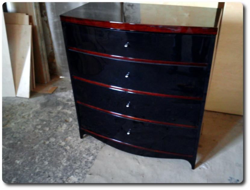 Gięta komoda z czterema szufladami w czarnym, wysokim połysku, z mahoniowymi akcentami, sfotografowana w warsztacie na tle materiałów budowlanych.