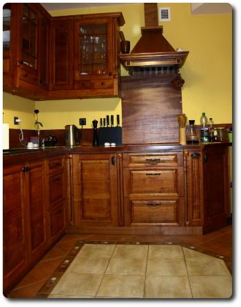 Rustykalna zabudowa kuchenna z ciemnego drewna z frezowanymi frontami, widoczny okap i płytki podłogowe w odcieniach beżu i brązu.