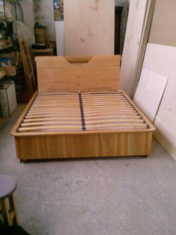 Drewniane łóżko z prostym zagłówkiem i stelażem listwowym, widoczne w warsztacie stolarskim, otoczone elementami drewnianymi i narzędziami.