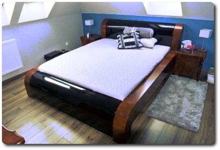 Łóżko z ciemnego drewna o zaokrąglonych kształtach w sypialni na poddaszu, z dwiema szafkami nocnymi i dywanikiem.