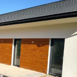 Nowoczesny dom jednorodzinny z elewacją w dwóch odcieniach: kremowy tynk i panele imitujące drewno, widoczne trzy okna przesuwne, szara dachówka ceramiczna i rynna spustowa.