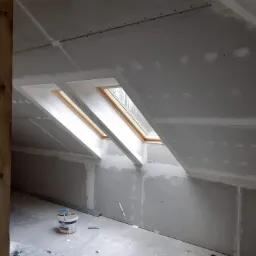 Poddasze w trakcie wykańczania płytami kartonowo-gipsowymi, widoczne dwa okna dachowe, drewniany słup i wiadro z masą szpachlową.