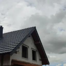 Nowy, grafitowy dach z blachodachówki na domu w trakcie budowy, widoczne okna poddasza i komin, pochmurne niebo.