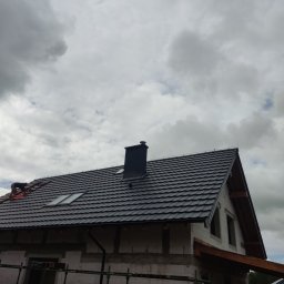 TOMASZ TOMCZAK TOM-BUD - Montaż czarnej dachówki na domu w stanie surowym otwartym, widoczny dekarz przy pracy, pochmurne niebo w tle.