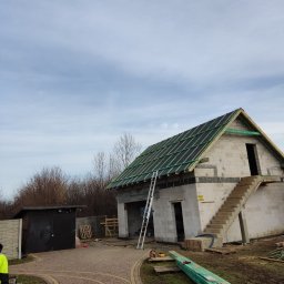TOMASZ TOMCZAK TOM-BUD - Budowa domu z częściowo położonym pokryciem dachowym z czarnych płyt i zielonych łat, widoczne rusztowanie i materiały budowlane na posesji.