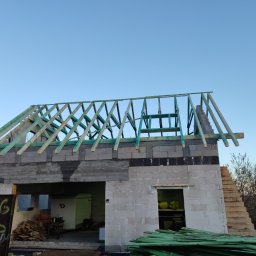 TOMASZ TOMCZAK TOM-BUD - Surowa konstrukcja domu z widoczną więźbą dachową z impregnowanego drewna, w tle błękitne niebo, elementy rusztowania i składowane deski na pierwszym planie.