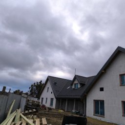 TOMASZ TOMCZAK TOM-BUD - Dwa domy jednorodzinne w stanie surowym zamkniętym z ciemną dachówką na tle pochmurnego nieba. Okna zabezpieczone folią, widoczne drewniane elementy konstrukcyjne i ogrodzenie.