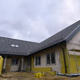 Dom jednorodzinny w trakcie ocieplania ścian zewnętrznych i montażu dachu z czarnej dachówki, widoczne rusztowania i materiały izolacyjne, pochmurne niebo w tle.