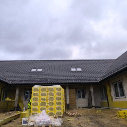 TOMASZ TOMCZAK TOM-BUD - Budowa domu jednorodzinnego: ułożona dachówka ceramiczna, okna dachowe, ocieplone ściany zewnętrzne żółtym styropianem, paleta z materiałem izolacyjnym pod ścianą, pochmurne niebo.