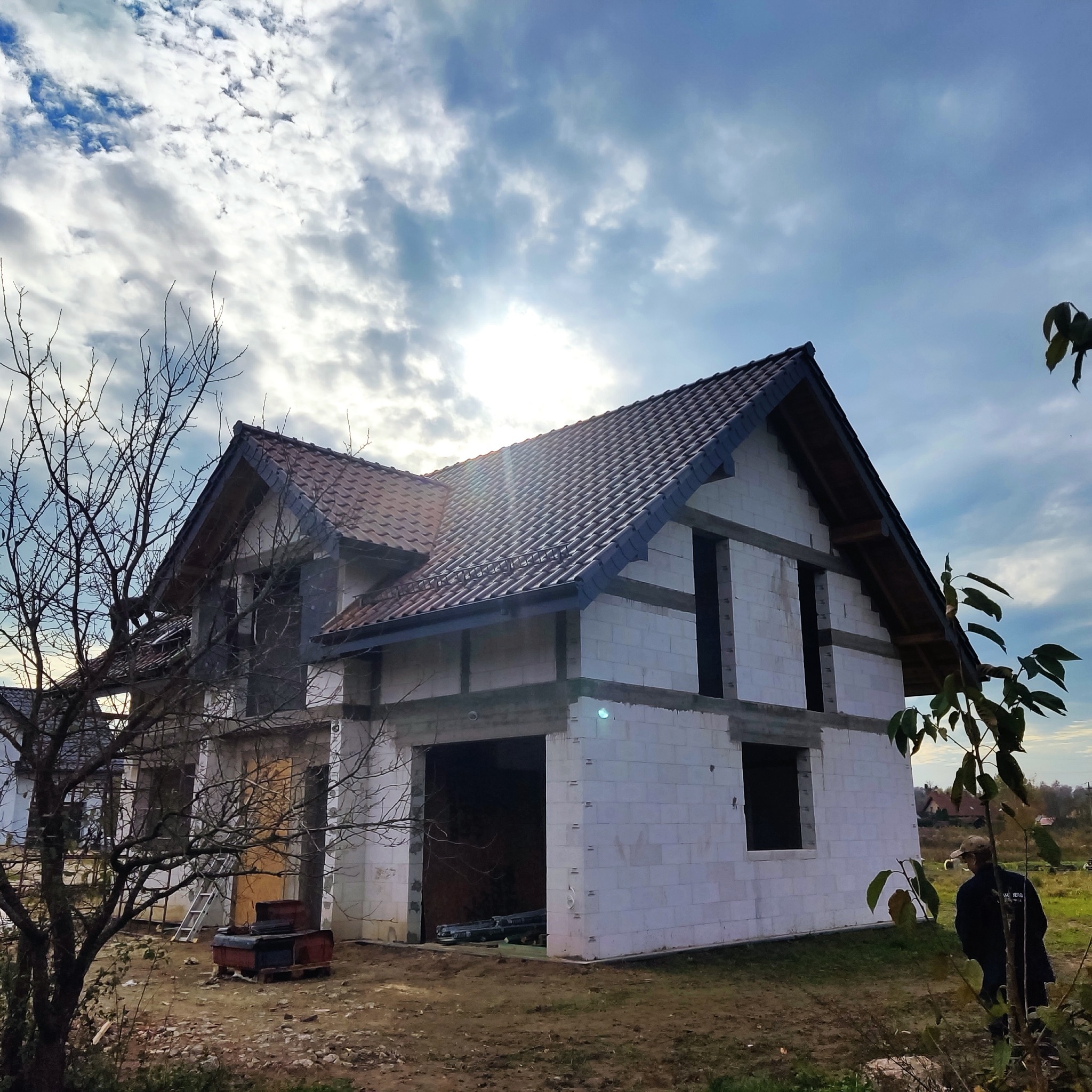 Dom jednorodzinny w trakcie budowy, z częściowo ukończonym dachem z brązowej dachówki, widoczne ściany z białego bloczka, otwarte okna, w tle błękitne niebo z chmurami, na pierwszym planie drzewo...