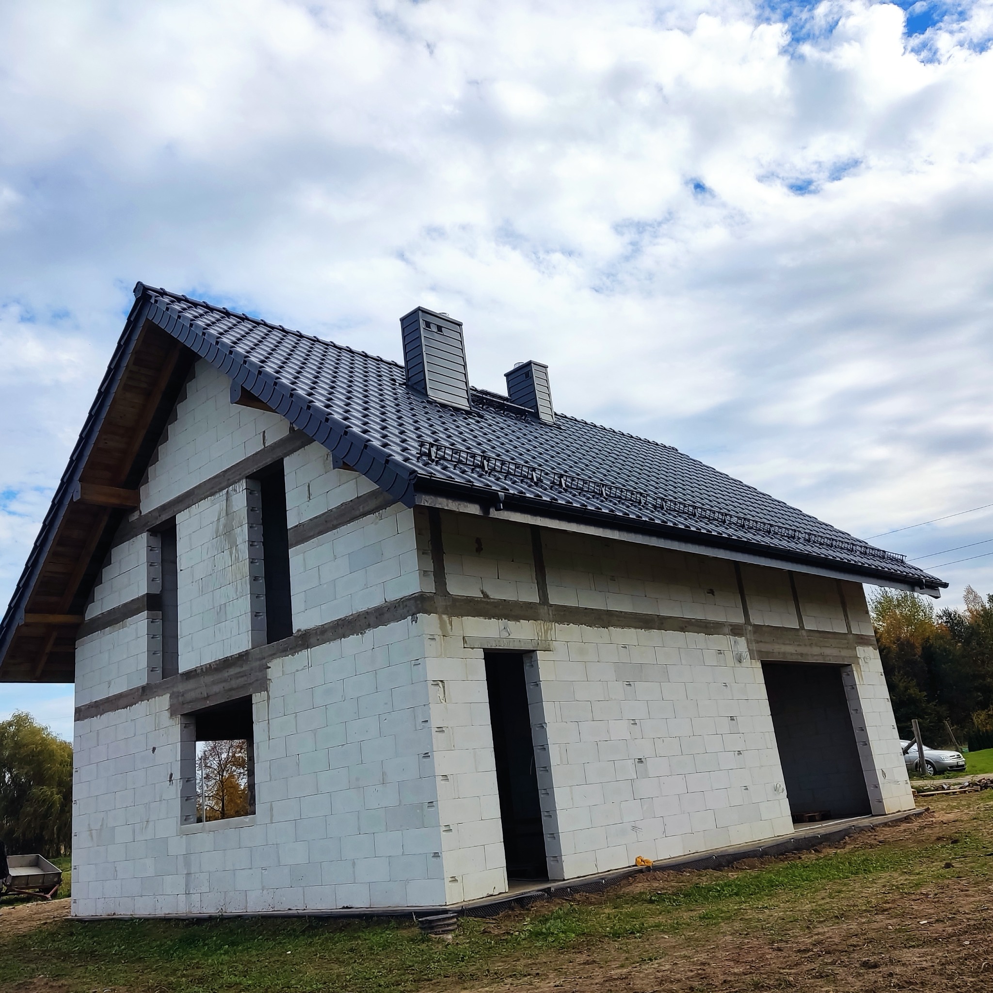 Surowy dom jednorodzinny w trakcie budowy, z czarną dachówką i kominami, widok na niezadaszony budynek z pustaków w pochmurny dzień.