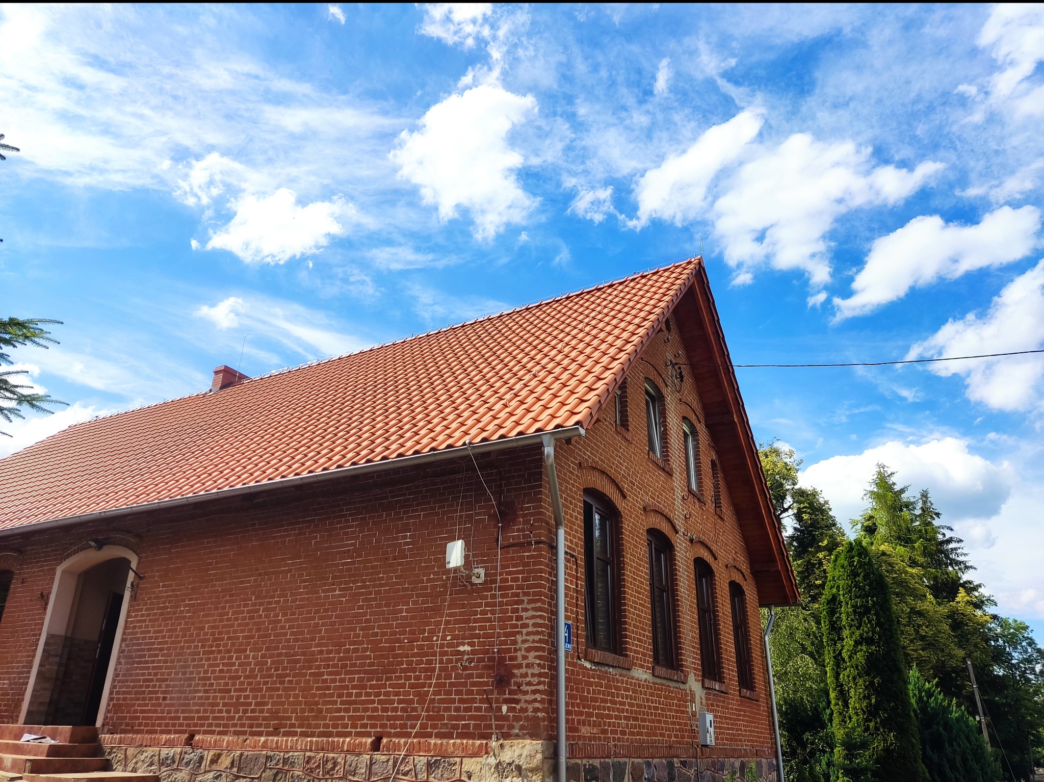Widok z dołu na budynek z czerwonej cegły z nową, czerwoną dachówką na tle błękitnego nieba z chmurami.