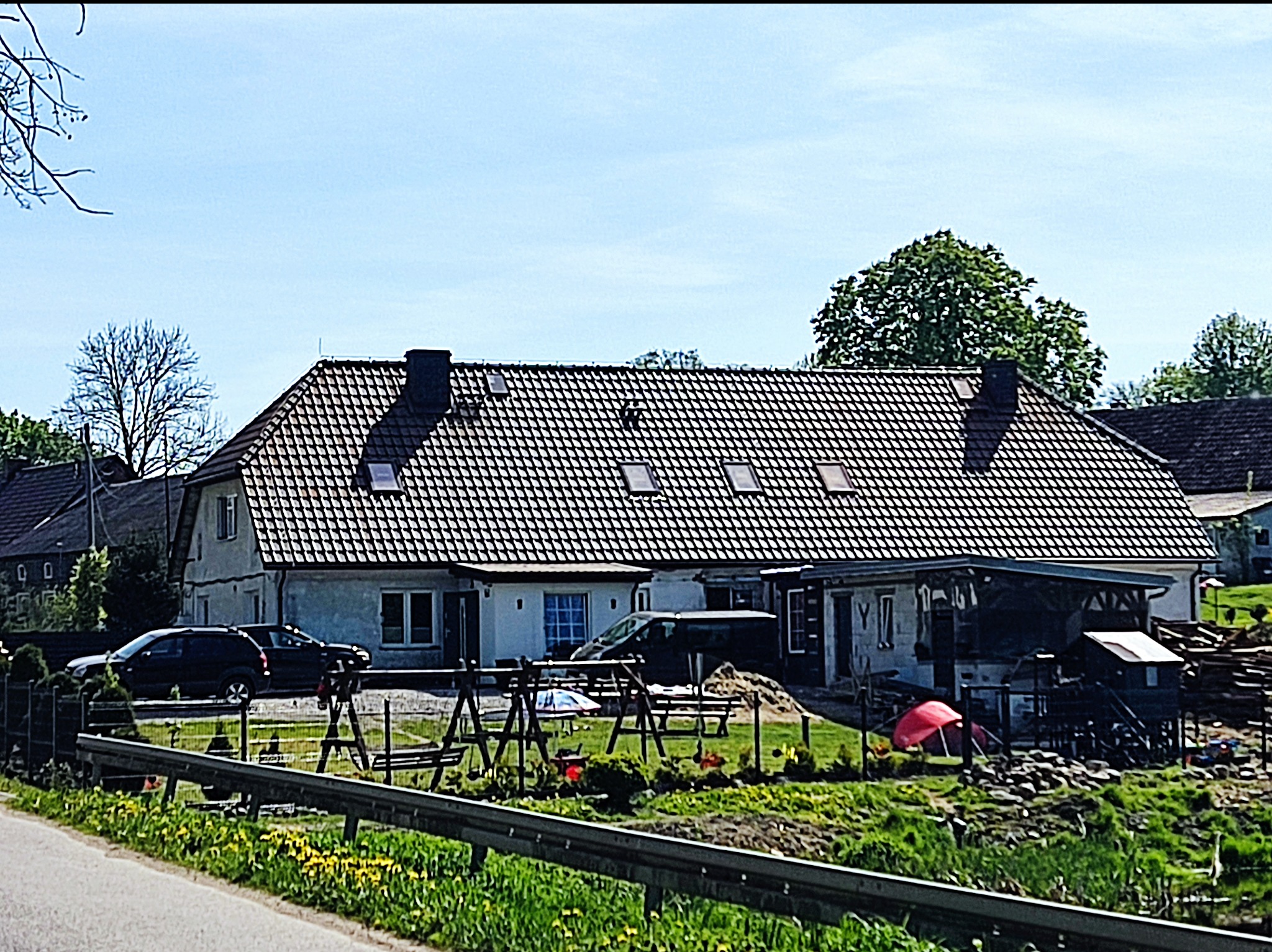 Dom jednorodzinny z ciemną dachówką karpiówką i oknami dachowymi, widoczny zza barierek, z elementami placu budowy w tle.