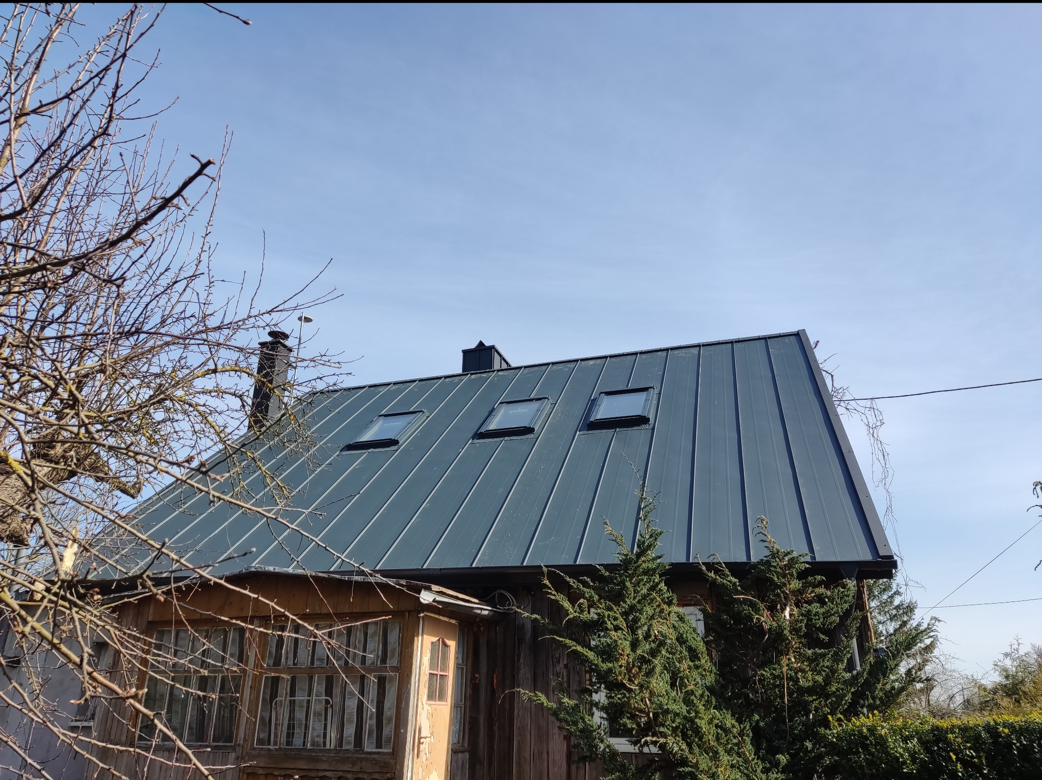 Dom z nowym, grafitowym dachem z blachy na rąbek stojący z trzema oknami dachowymi, widoczny komin i fragment drewnianej elewacji z oknami i drzwiami, na pierwszym planie gałęzie drzewa i iglaki.