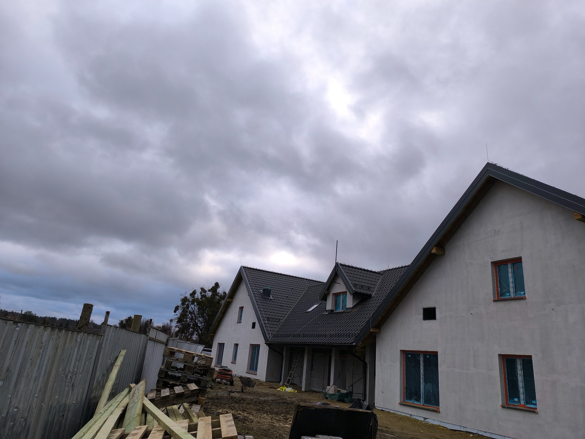 Dwa domy jednorodzinne w stanie surowym zamkniętym z ciemną dachówką na tle pochmurnego nieba. Okna zabezpieczone folią, widoczne drewniane elementy konstrukcyjne i ogrodzenie.