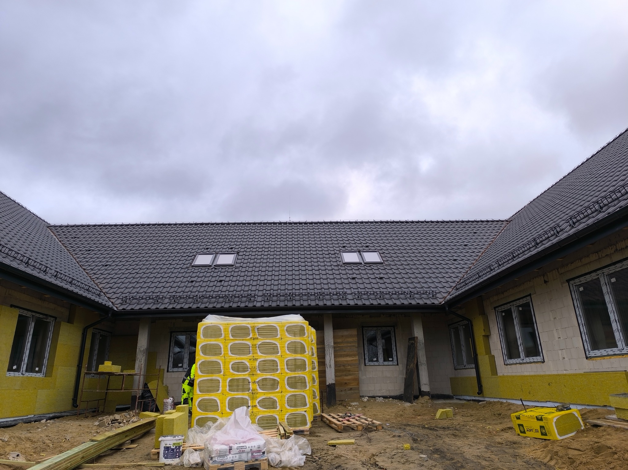 Budowa domu jednorodzinnego: ułożona dachówka ceramiczna, okna dachowe, ocieplone ściany zewnętrzne żółtym styropianem, paleta z materiałem izolacyjnym pod ścianą, pochmurne niebo.