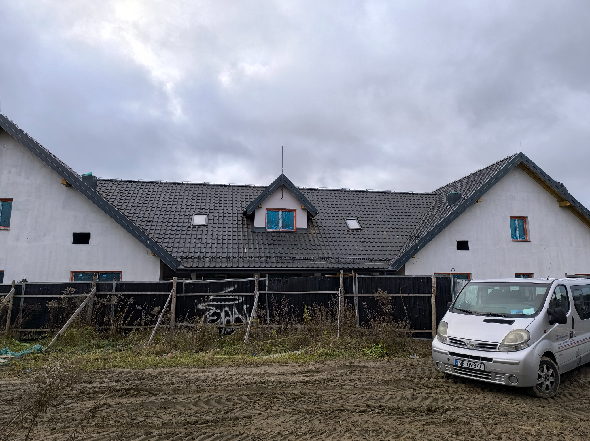 Nowy dom jednorodzinny z ciemną dachówką karpiówką, widoczne obróbki blacharskie, ogrodzenie z graffiti i srebrny bus firmy budowlanej na błotnistym terenie.