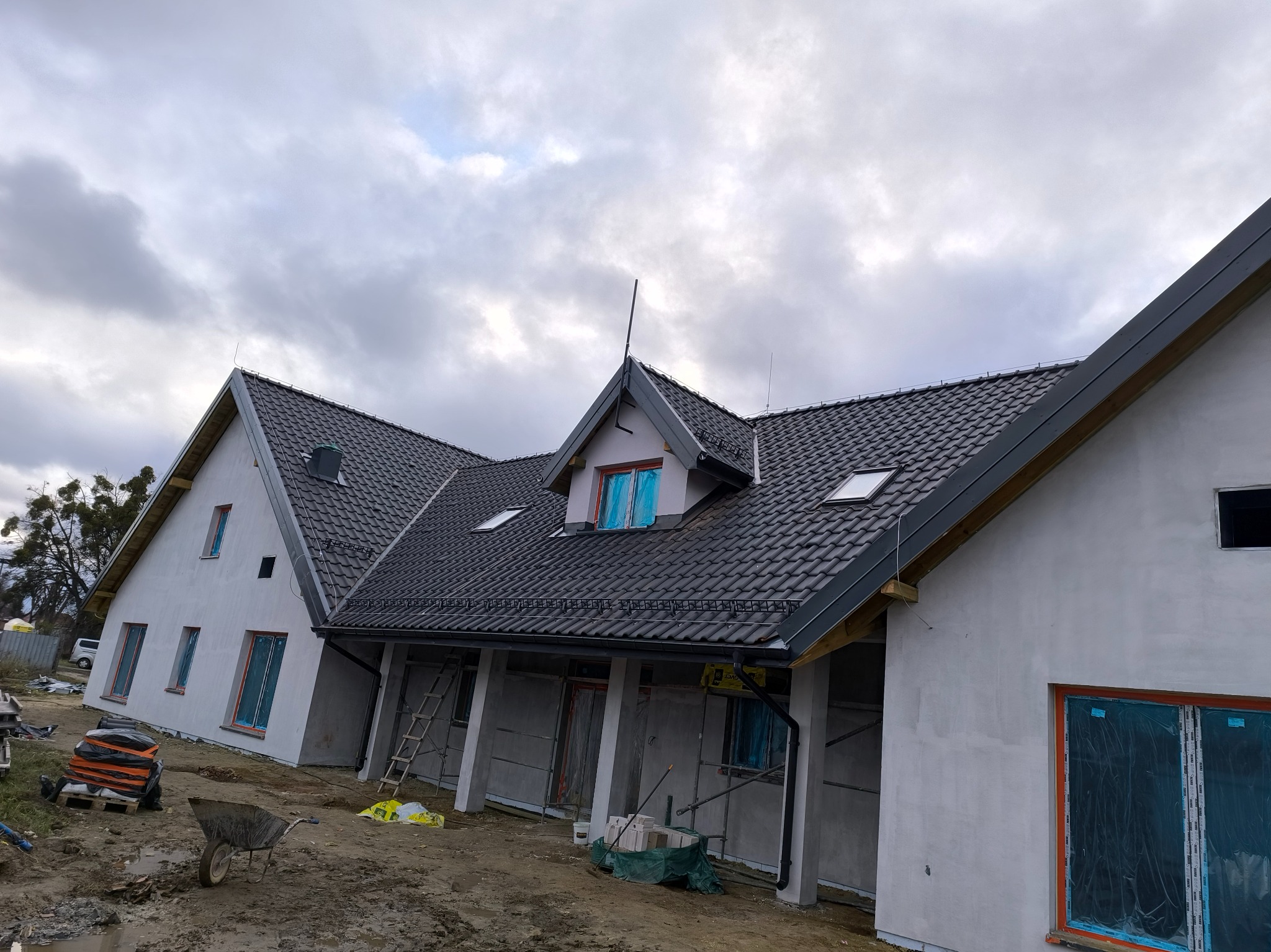 Nowy dom w trakcie budowy z grafitową dachówką, zabezpieczone okna folią, widoczny wózek budowlany i drabina oparta o ścianę.