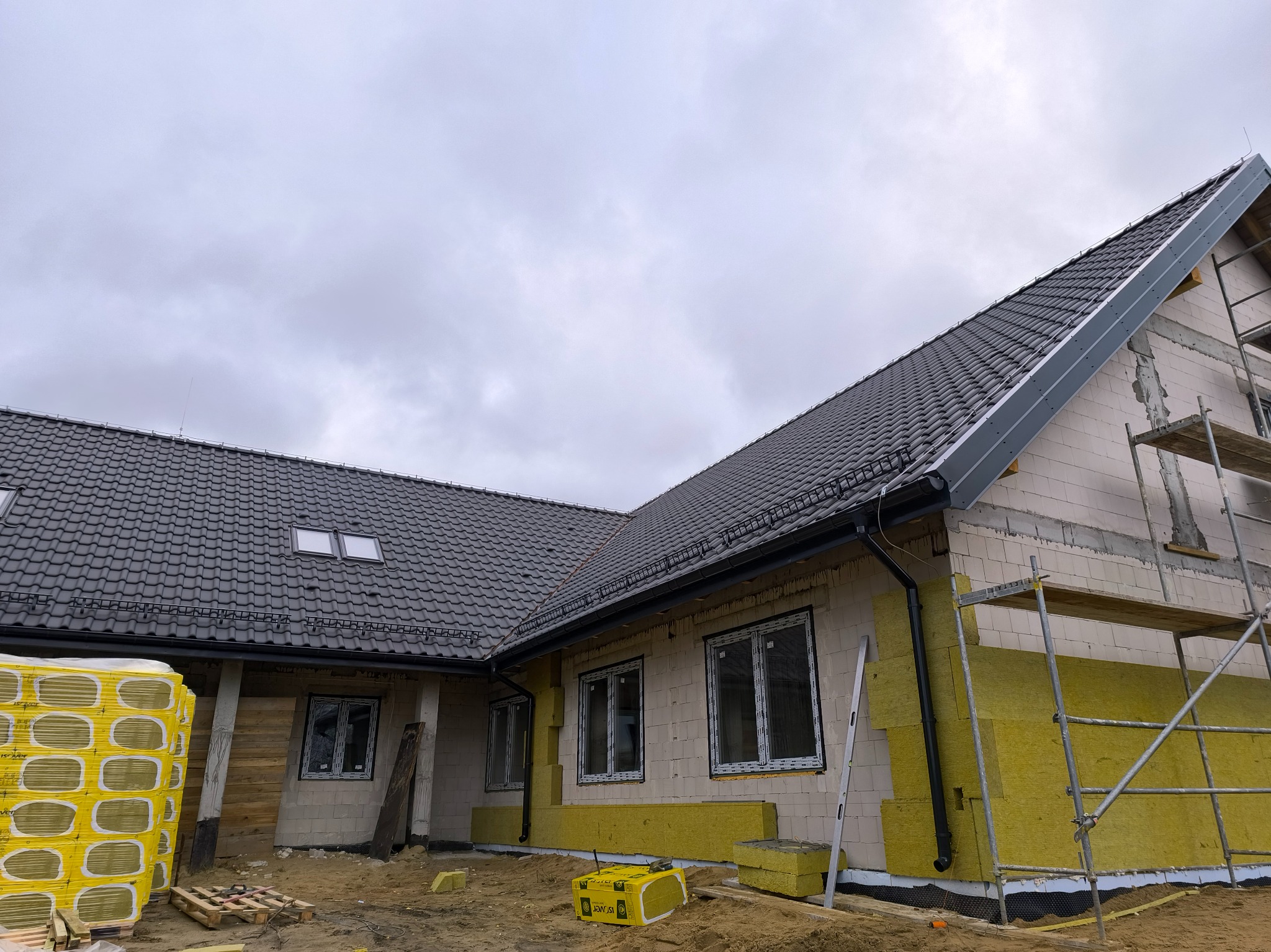Dom w trakcie budowy z szarą dachówką, ocieplony wełną mineralną, widoczne rusztowania i materiały budowlane na ziemi, pochmurne niebo.