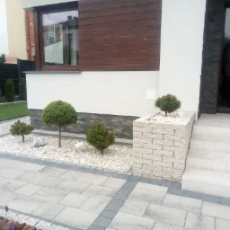 Nowoczesna aranżacja przestrzeni przed domem z kostką brukową, ozdobnymi kamieniami i niskimi drzewkami kulistymi w donicach z jasnej cegły, oraz schodami prowadzącymi do drzwi wejściowych.