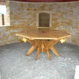 Ośmiokątny, drewniany stół z dekoracyjnym wzorem na blacie, umieszczony wewnątrz murowanej konstrukcji z kamienia, z oświetleniem punktowym w podłodze z kostki brukowej.