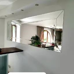 Prostokątne lustro z fazowanymi krawędziami, zamontowane na jasnoszarej ścianie w nowoczesnym wnętrzu, odbijające widok na salon z roślinami i oknem.