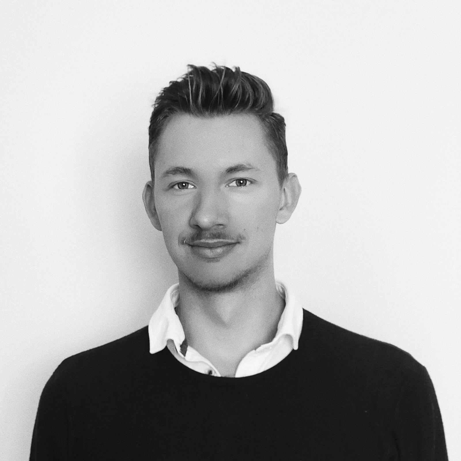 Portret młodego mężczyzny w czarno-białej tonacji, ubranego w ciemny sweter i białą koszulę z kołnierzykiem, na tle jednolitej, jasnej ściany.