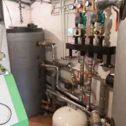 Instalacja kotłowni z dwoma zbiornikami akumulacyjnymi, licznymi rurami i zaworami, widok na brązowe płytki podłogowe i fragment zielonej obudowy urządzenia.