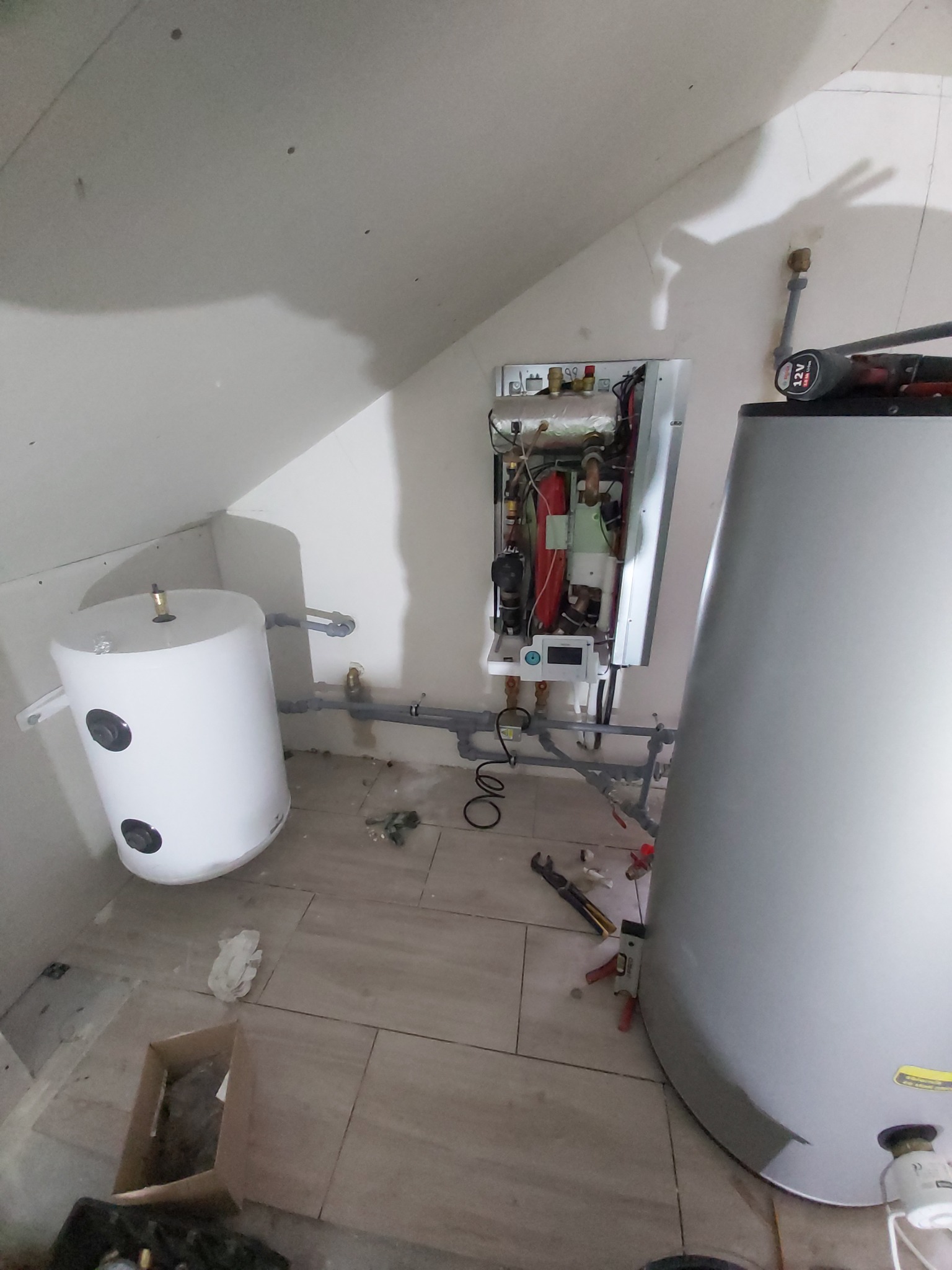 Biały zbiornik akumulacyjny ciepłej wody i rozłożony moduł centrali wentylacyjnej z odzyskiem ciepła, widoczne rury, narzędzia i karton na podłodze w trakcie montażu.