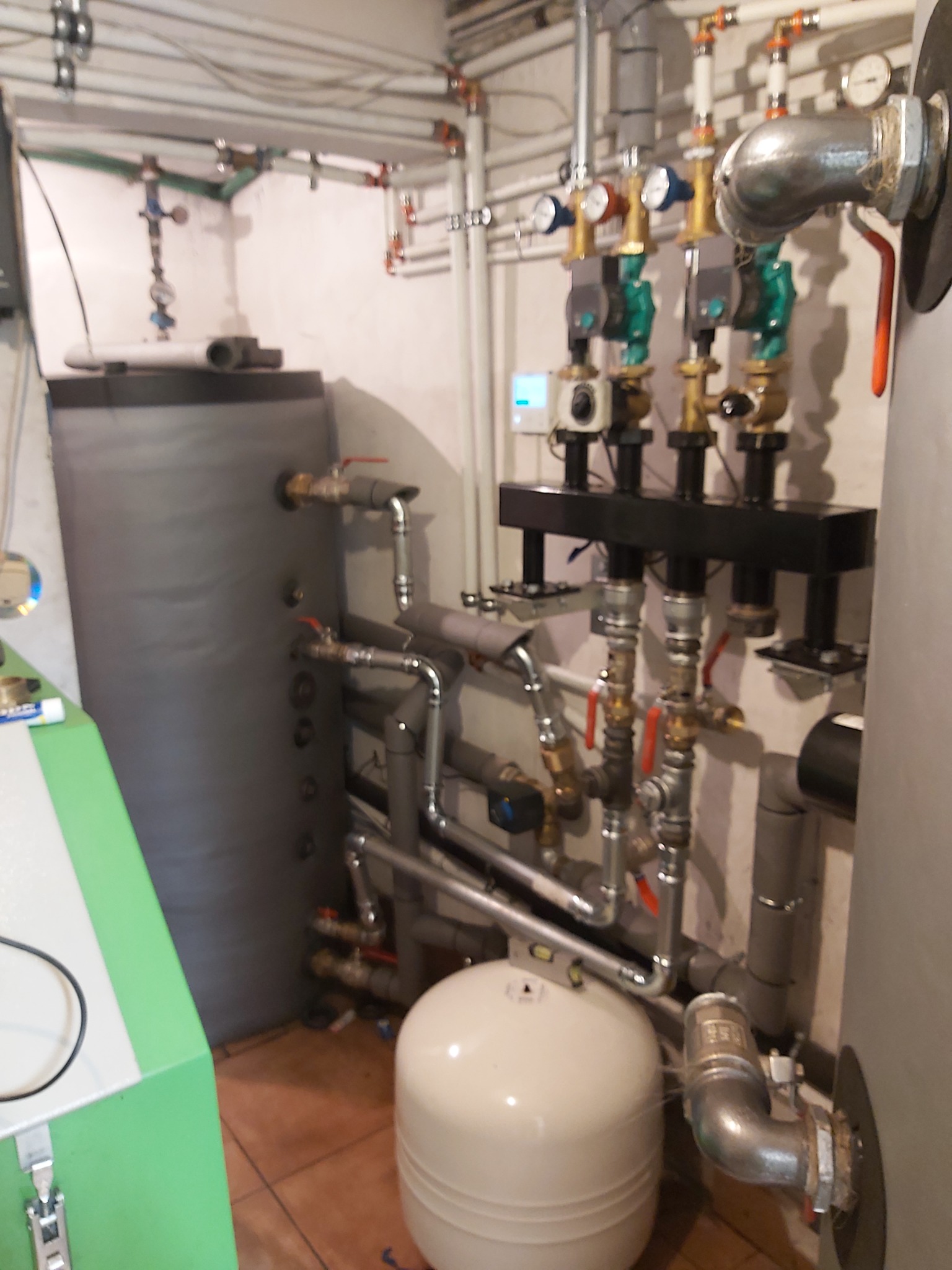 Instalacja kotłowni z dwoma zbiornikami akumulacyjnymi, licznymi rurami i zaworami, widok na brązowe płytki podłogowe i fragment zielonej obudowy urządzenia.