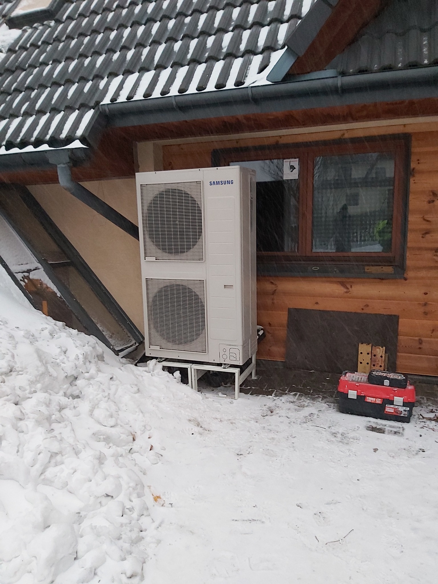 Biała pompa ciepła Samsung zamontowana na zewnątrz drewnianego domu w zimowej scenerii, obok widoczna skrzynka z narzędziami.