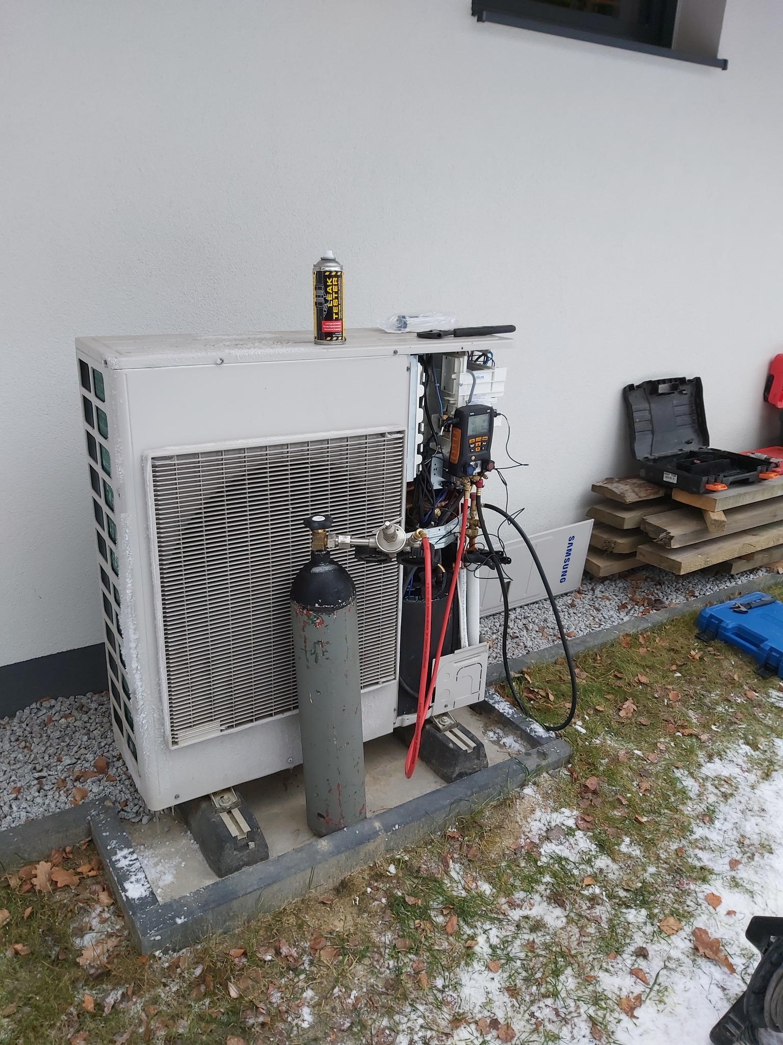 Zewnętrzna jednostka klimatyzacji Samsung z podłączonymi manometrami i butlą z gazem chłodniczym, na górze puszka Leak Tester, częściowo pokryta śniegiem, na zewnątrz budynku.