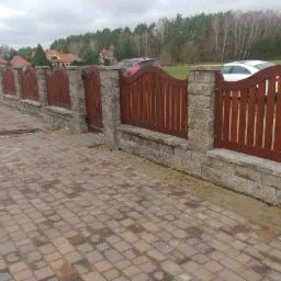 Ogrodzenie z drewnianymi panelami i kamiennymi słupkami, z bramą, na posesji z kostką brukową. Widoczne domy w tle i drzewa na horyzoncie.