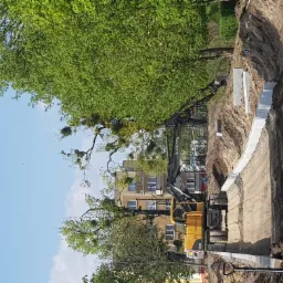 Żółta koparka Volvo wykonuje wykop w ziemi, widoczny betonowy fundament w wykopie, drzewa w tle