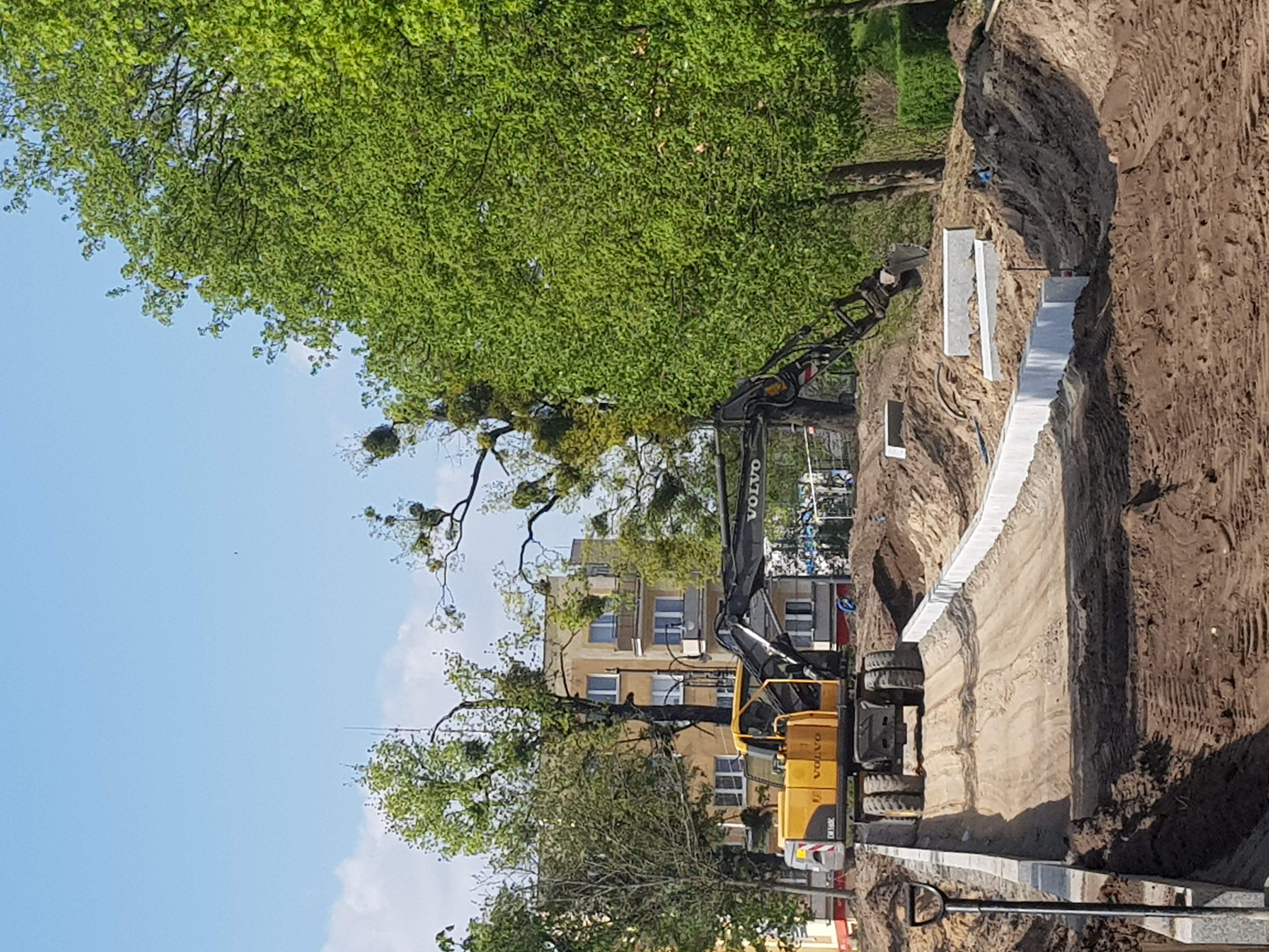 Żółta koparka Volvo wykonuje wykop w ziemi, widoczny betonowy fundament w wykopie, drzewa w tle