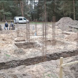 Łukasz Trusiak Zakład Ogólnobudowlany - Przygotowanie fundamentów pod budowę domu: wykopy, zbrojenie stalowe, szalunki w ziemi. W tle ekipa budowlana i samochód dostawczy na skraju lasu.
