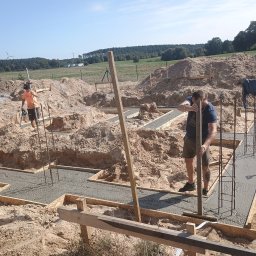 Łukasz Trusiak Zakład Ogólnobudowlany - Wylewanie fundamentów pod dom na piaszczystym terenie. Widoczne drewniane szalunki i stalowe zbrojenia. Dwóch pracowników budowlanych w trakcie pracy.