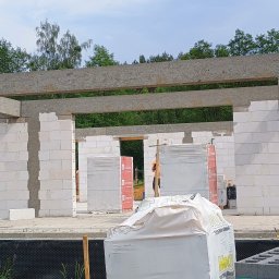 Łukasz Trusiak Zakład Ogólnobudowlany - Budowa domu w trakcie: białe bloczki, betonowe nadproża, materiały budowlane na placu. Widoczny zarys ścian i otwory okienne. W tle zieleń drzew.