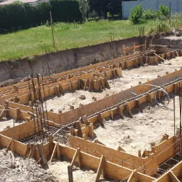 Szczegółowy widok z góry na szalunek fundamentów domu zbrojonego stalą, przygotowany do zalania betonem, na tle zieleni i betonowego ogrodzenia.