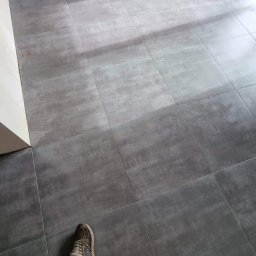 Łukasz Trusiak Zakład Ogólnobudowlany - Szare płytki podłogowe ułożone w kwadraty, widoczne nogi w butach sportowych. Jasne światło pada na podłogę, podkreślając teksturę płytek.