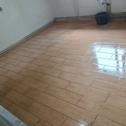 Łukasz Trusiak Zakład Ogólnobudowlany - Ukończona podłoga z płytek imitujących drewno w jasnym odcieniu, świeżo po ułożeniu, z widocznym wiaderkiem i rurami przy ścianie. Ujęcie z niskiego kąta.