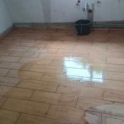 Łukasz Trusiak Zakład Ogólnobudowlany - Ułożona podłoga z płytek imitujących drewno w jasnym odcieniu, widoczne rury przy ścianie i czarne wiadro. Podłoga mokra, odbija światło.