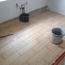 Łukasz Trusiak Zakład Ogólnobudowlany - Ułożone, podłużne płytki podłogowe imitujące drewno w łazience. Widoczne narzędzia glazurnicze, wiadro i poziomica. Częściowo wykończona podłoga w jasnych barwach.