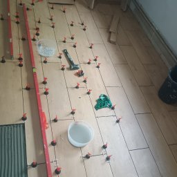 Łukasz Trusiak Zakład Ogólnobudowlany - Układanie płytek imitujących drewno na podłodze z użyciem systemu poziomowania. Widoczny poziomica, młotek gumowy i pojemnik z klinami.