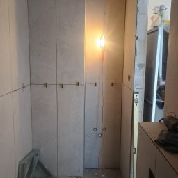 Łukasz Trusiak Zakład Ogólnobudowlany - Trwające prace glazurnicze w łazience: ściany pokryte jasnymi, wielkoformatowymi płytkami z widocznymi klipsami poziomującymi, oświetlone tymczasową lampą roboczą.