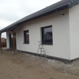 Nowo otynkowany, parterowy dom z ciemną dachówką i drewnianą konstrukcją zadaszenia tarasu, z drabinką opartą o ścianę i paletami w tle.