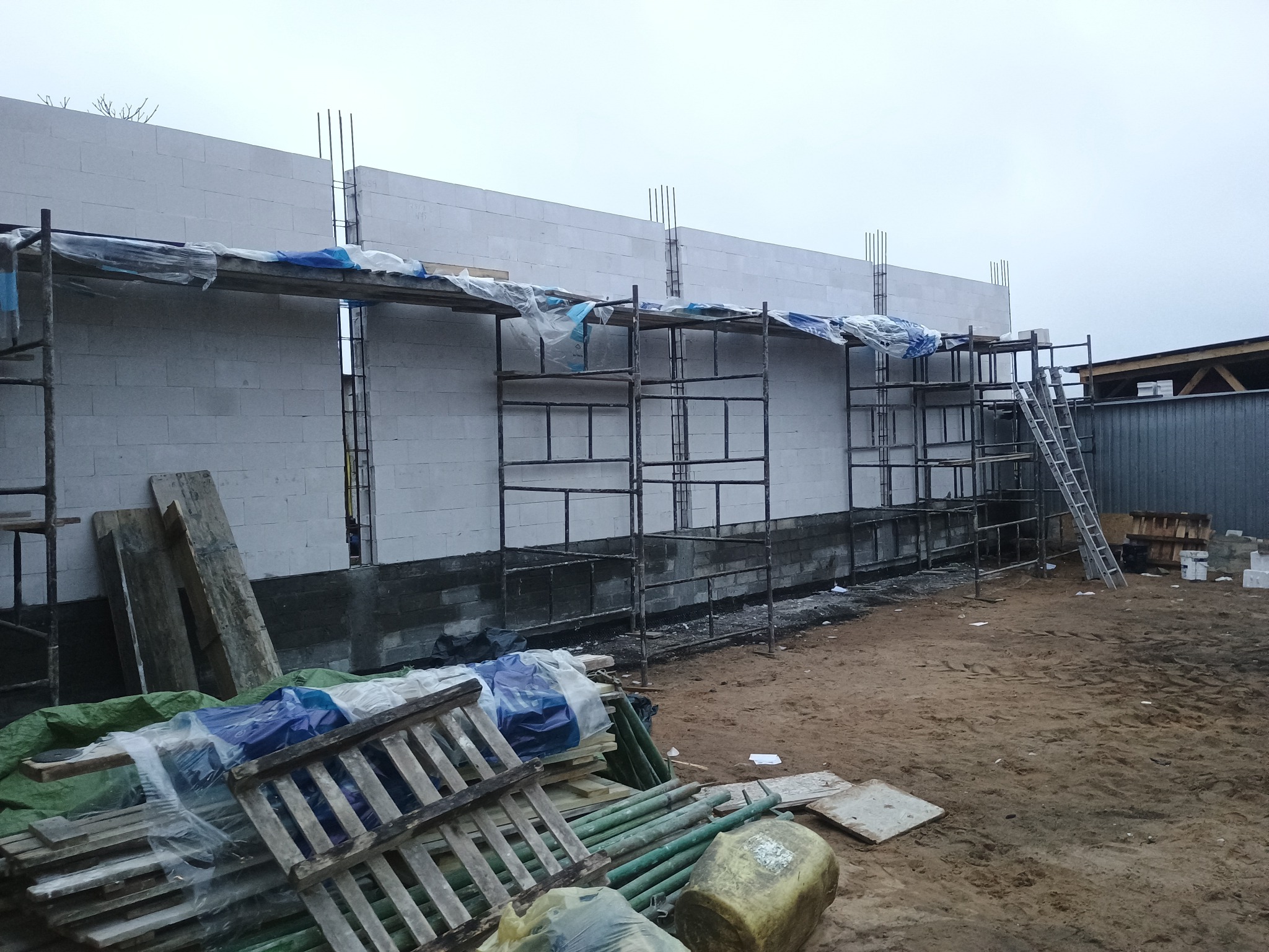 Wznoszenie ściany z bloczków betonu komórkowego na fundamencie. Widoczne rusztowania, wystające pręty zbrojeniowe i materiały budowlane na placu budowy.