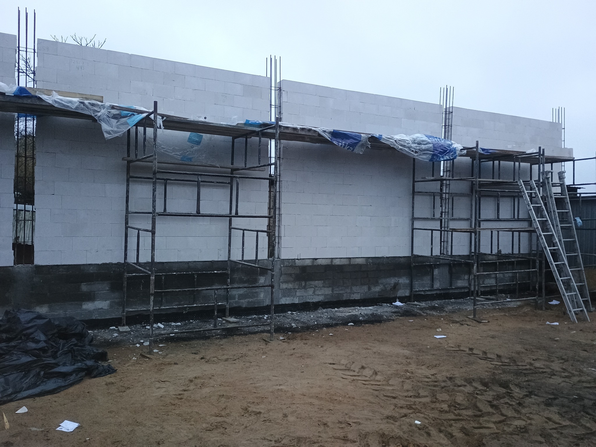 Wznoszenie ścian z białych bloczków na fundamencie. Widoczne rusztowania, wystające pręty zbrojeniowe. Etap budowy domu w pochmurny dzień.