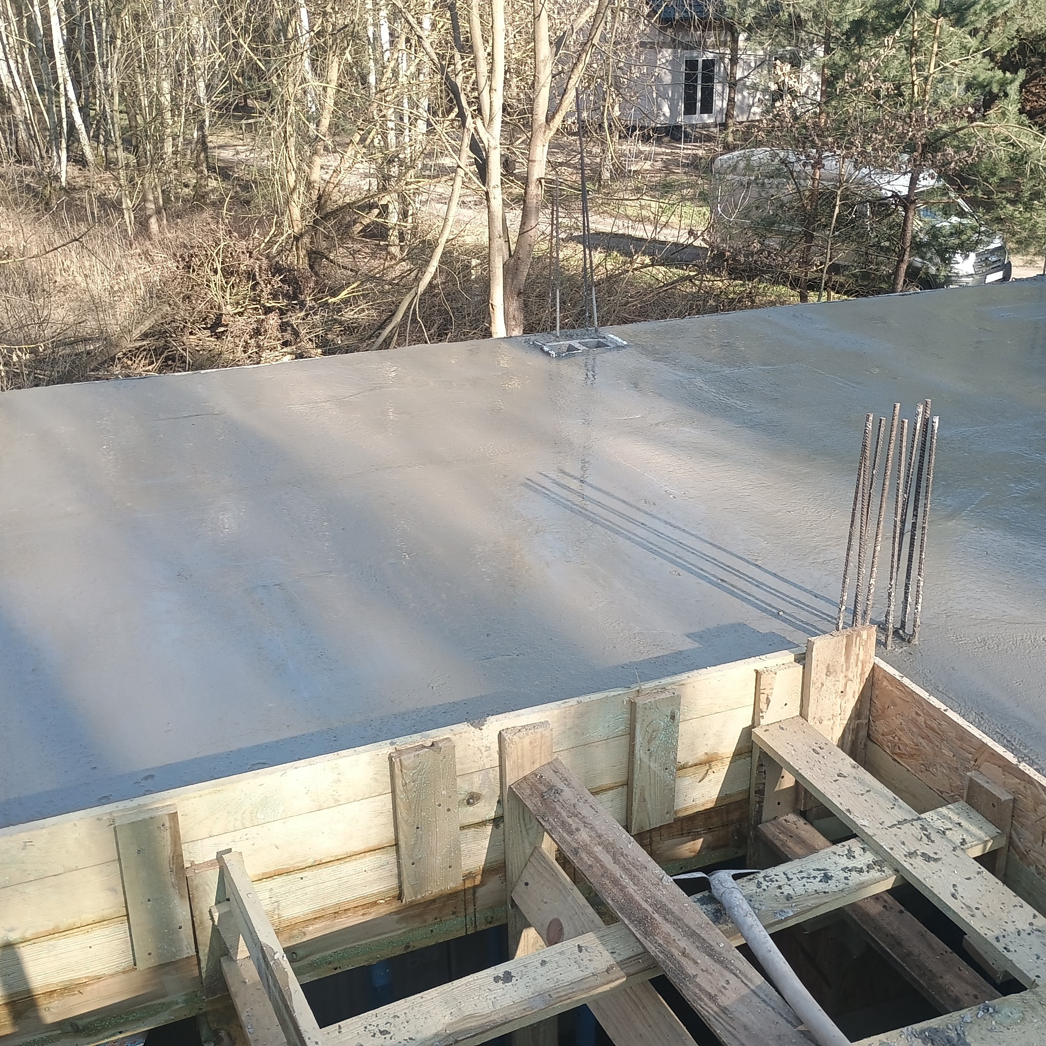 Wylany beton na stropie z wystającymi prętami zbrojeniowymi i drewnianym szalunkiem. W tle drzewa i budynek. Widok z góry, ostre światło słoneczne.