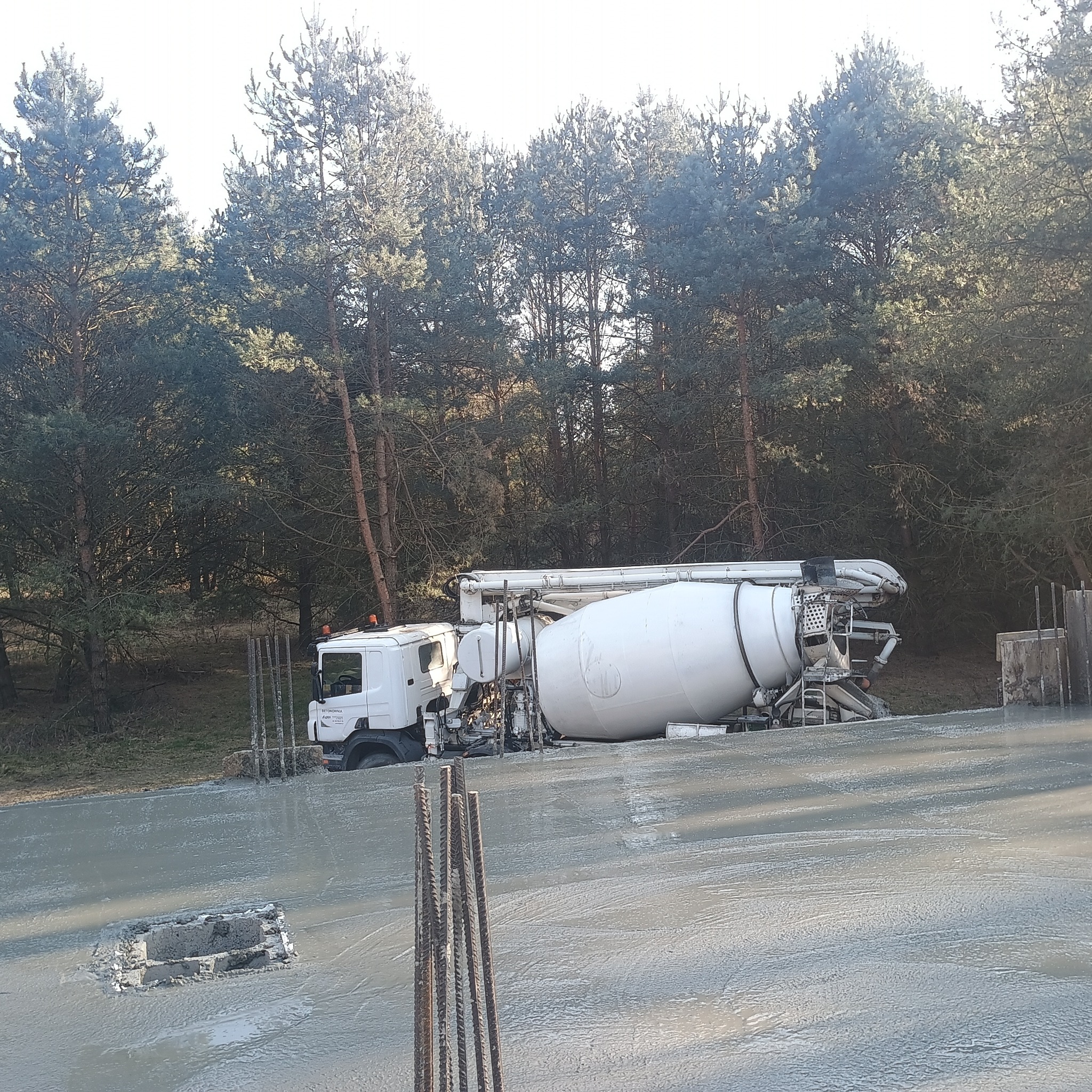 Fundament domu w trakcie wylewania betonu, widoczny mikser do betonu i wystające pręty zbrojeniowe na tle lasu.